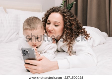 Happy family mom and cute little baby son laughing use smartphone funny face mask app look at cell screen, mom with baby take selfie watch cartoons make video call on mobile sit on sofa at home