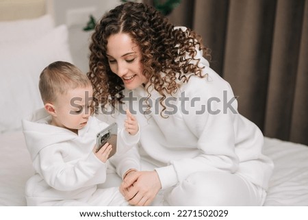 Happy family mom and cute little baby son laughing use smartphone funny face mask app look at cell screen, mom with baby take selfie watch cartoons make video call on mobile sit on sofa at home