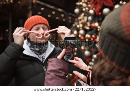 Positive couple in love wearing hats and winter clothes outdoors at big christmas tree background taking funny pictures of each others using smart phone