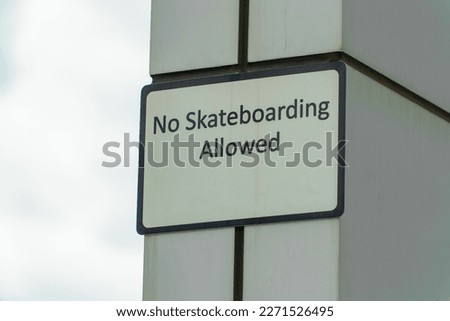Sign that says no skateboarding allowed near a school or in a public area of neighborhood park or city downtown streets. Black and white sign with gray cement wall and cloudy sky background.