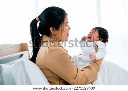 Asian mother hold crying newborn baby and stay on bed in bedroom with day light and she look care and love with baby.
