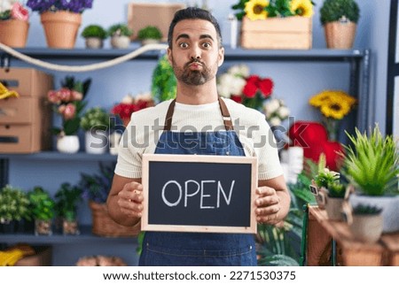 Hispanic man with beard working at florist holding open sign puffing cheeks with funny face. mouth inflated with air, catching air. 