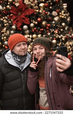 Positive couple in love wearing hats and winter clothes outdoors in city street at big christmas tree background doing selfie photo by smart phone. Vertical banner