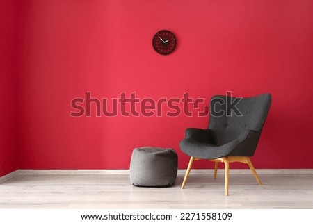 Stylish grey armchair, pouf and clock near red wall