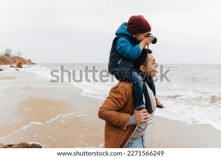 Cute little boy sitting on his father's shoulders and taking pictures while spending time together on beautiful beach
