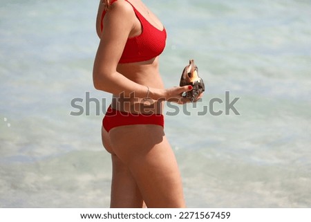 Slim woman in red swimsuit standing in sea water with seashell in hand. Beach vacation on Caribbean islands