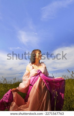 Beautiful girl in a lush pink ball gown in green field during blooming of flowers and blue sky on background. Model posing on nature landscape as princess from fary tale
