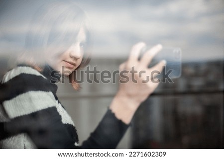 Influencer teenage girl in the city by the window, using a smartphone. Background reflection of the sky on the window. Side view, bright, texture.