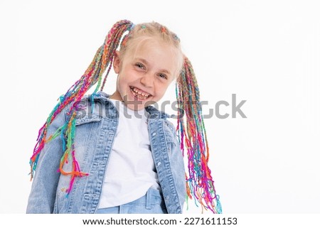 Delighted blonde daughter with colorful braids smiling at camera wearing denim costume standing on white background in studio posing for camera. Kid in good mood laughing spending time.