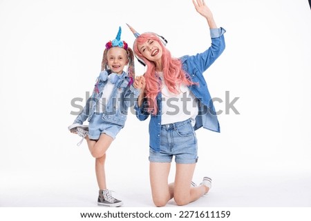 Happy adorable mother with pink wig and unicorn headband hugging preschooler daughter with braids standing on white background in studio isolaed in denim clothes having fun posing fo camera.