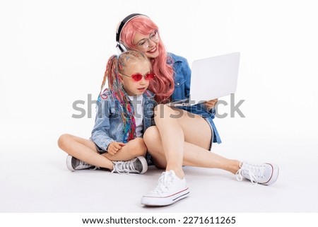 Adorable curious daughter wearing pink sunglasses and funny hairstyle looking at computer screen watching movies with young mother wearing pink wig and earphones holding laptop in arms.