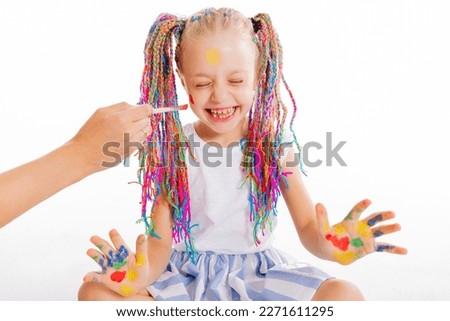 School girl siitting on white background in studio with colorful pigtails braids closing eyes laughing having fun with mother who paints on cheeks using painting brushes small kid with colorful palm.
