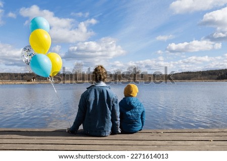 unrecognizable little boy and woman sit with their backs on the shore of a lake with blue and yellow balloons. Family, refugees, unity, support, patriotism, nostalgia. stand with Ukraine