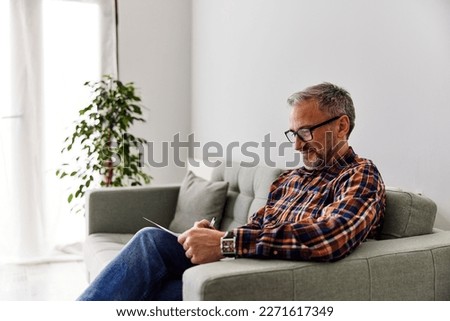 A smiling man with glasses on sitting on the cozy couch at home, holding a piece of paper, writing something. Royalty-Free Stock Photo #2271617349