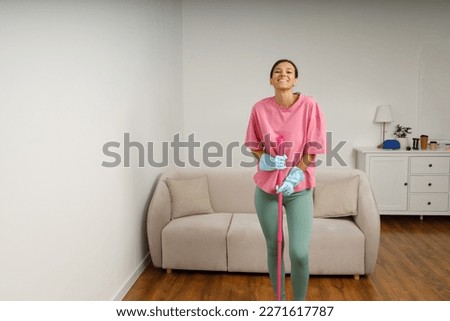 Full-size picture of happy smiling girl doing house cleaning. Washing floors. Cute young woman in casual clothes and rubber gloves wipes the floor with mop in the living room.