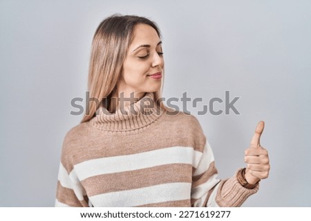 Young blonde woman wearing turtleneck sweater over isolated background looking proud, smiling doing thumbs up gesture to the side 