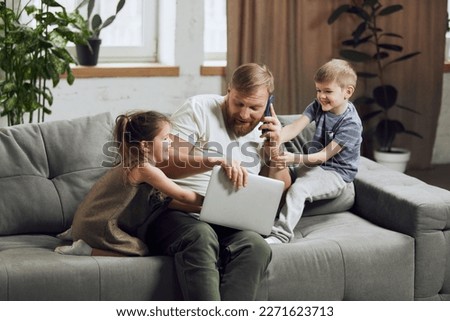 Man, father sitting on couch, talking on phone and working on laptop in living room. Kids bothering and playing with him. Concept of fatherhood, childhood, family, freelance job, remote work Royalty-Free Stock Photo #2271623713