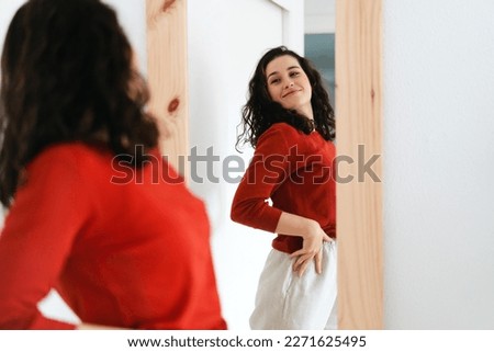  Young beautiful self confident woman in red sweater looking in the mirror proud of herself Royalty-Free Stock Photo #2271625495