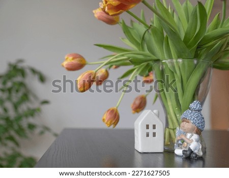 Figure of a girl with flowers and a house near a vase with tulips against the background of a white wall.