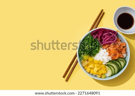Poke bowl with rice, salmon,cucumber,mango,onion,wakame salad, poppy seeds ands sunflowers seeds on yellow background. Top view. Copy space