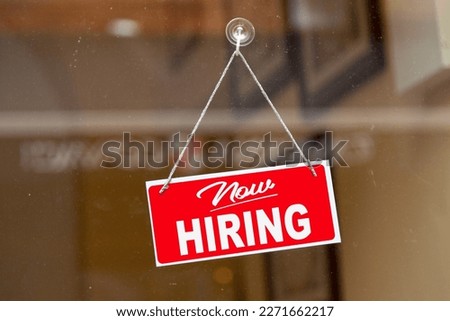 Red sign hanging at the glass door of a shop saying: "Now hiring". Royalty-Free Stock Photo #2271662217