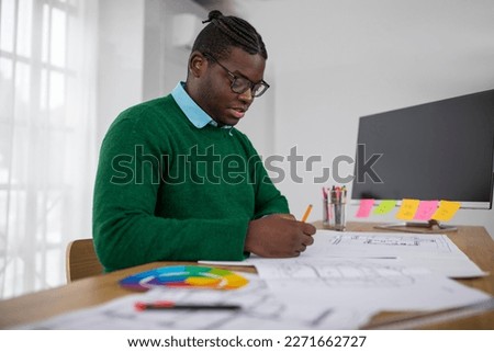 Side View Of Black Designer Man Drawing Sitting At Desk Covered With Papers Working Via Pencil And Ruler Creating Plan In Office. Modern Architecture And Design Career Concept