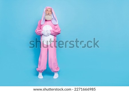Full length photo of cheerful man dressed pink rabbit costume look empty space hold hands together isolated on blue color background