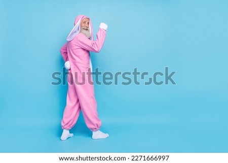Side profile full size photo of cheerful elderly pensioner wear pink bunny costume demonstrating biceps isolated on blue color background
