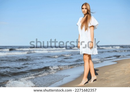 Happy smiling beautiful woman is walking on the ocean beach in a white summer dress.