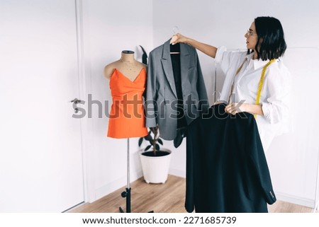 Young stylish female stylist in glasses picking jacket for orange top put on mannequin while creating outfit in own studio Royalty-Free Stock Photo #2271685739