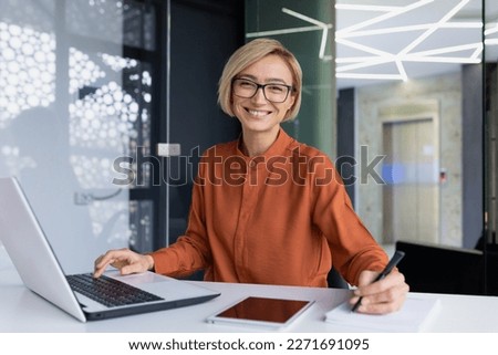Successful and happy business woman looking at camera sitting inside office working with documents, portrait of satisfied woman writing in notebook and working using laptop at work. Royalty-Free Stock Photo #2271691095