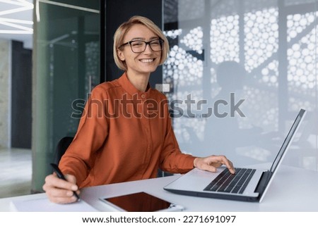 Successful and happy business woman looking at camera sitting inside office working with documents, portrait of satisfied woman writing in notebook and working using laptop at work. Royalty-Free Stock Photo #2271691097