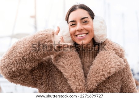 Young woman wearing winter muffs at outdoors making phone gesture. Call me back sign