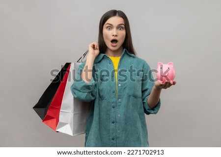 Amazed astonished woman with dark hair holding shopping bags and piggy bank, cashback from buying purchases, wearing casual style jacket. Indoor studio shot isolated on gray background.