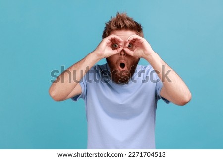 Portrait of wondered shocked bearded man making binoculars gesture with hands and looking amazed at camera, keeps mouth opened. Indoor studio shot isolated on blue background.