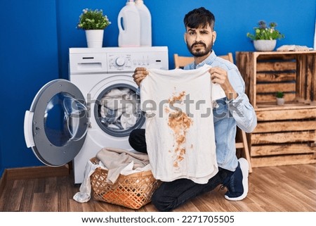 Young hispanic man with beard holding clean white t shirt and t shirt with dirty stain clueless and confused expression. doubt concept.  Royalty-Free Stock Photo #2271705505