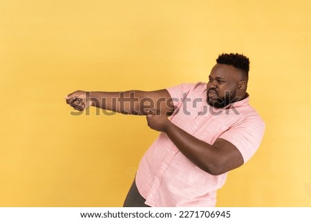 Portrait of persistent man with beard wearing pink shirt pretending to pull, , holding heavy virtual burden with expression of great effort. Indoor studio shot isolated on yellow background. Royalty-Free Stock Photo #2271706945