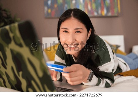 Portrait photo of delighted asian kazachstan girl in casual clothes outfit smiling at camera while laying on bed in front of laptop computer doing purchase shopping online holdin credit card in hands.