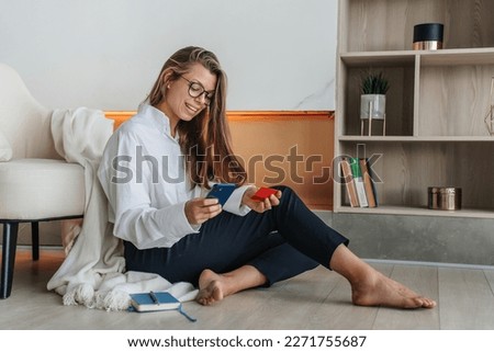 Cheerful hispanic young adult woman in white shirt and dark blue pants sits on floor at living room holds phone and credit card makes order via internet against fireplace and book shelfs on background