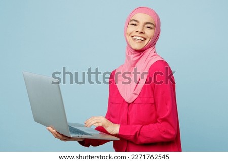 Side view young arabian muslim IT woman wear pink abaya hijab hold use work on laptop pc computer isolated on plain pastel light blue cyan background studio portrait People uae islam religious concept