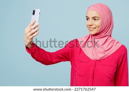 Young smiling arabian muslim woman wear pink abaya hijab doing selfie shot on mobile cell phone isolated on plain pastel light blue cyan background studio portrait. People uae islam religious concept