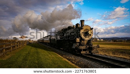 A View of a Classic Steam Passenger Train Approaching, With American Flags Attached to a Fence on a Sunny Autumn Day Royalty-Free Stock Photo #2271763801