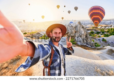 Happy bearded guy traveling blogger with victory sign takes selfie photo in Nevsehir, Goreme. Beautiful destination with flying hot air balloons in Anatolia, Kapadokya