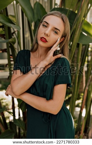 Tender stylish woman with blond hair and red lips is touching her check and looking at camera while posing during photoshoot on background of exotic plants 