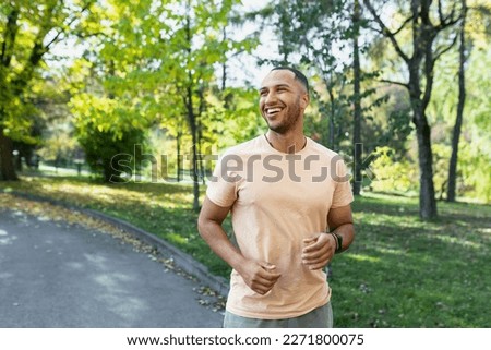 Cheerful and successful hispanic man jogging in the park, man running on a sunny day, smiling and happy having an outdoor activity. Royalty-Free Stock Photo #2271800075