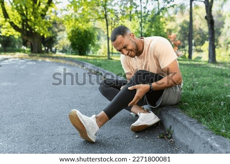 Runner injured his leg while jogging in the park near the trees, hispanic man has muscle spasm massaging his knee, man is running and exercising fitness on a sunny day. Royalty-Free Stock Photo #2271800081