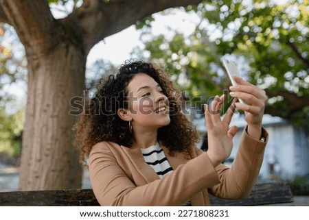 pretty curly smiling woman walking in city street in stylish jacket, using smartphone, taking selfie picture, making photo