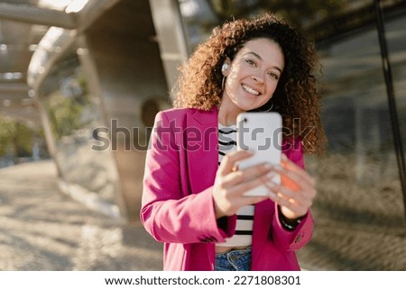 pretty curly woman in city street in stylish pink jacket, student education, using smartphone, taking selfie picture, making photo