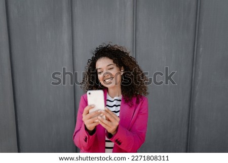 pretty curly smiling woman walking in city street in stylish pink jacket, using smartphone, taking selfie picture, making photo