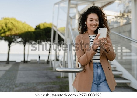 pretty curly smiling woman walking in city street in stylish jacket, using smartphone, taking selfie picture, making photo